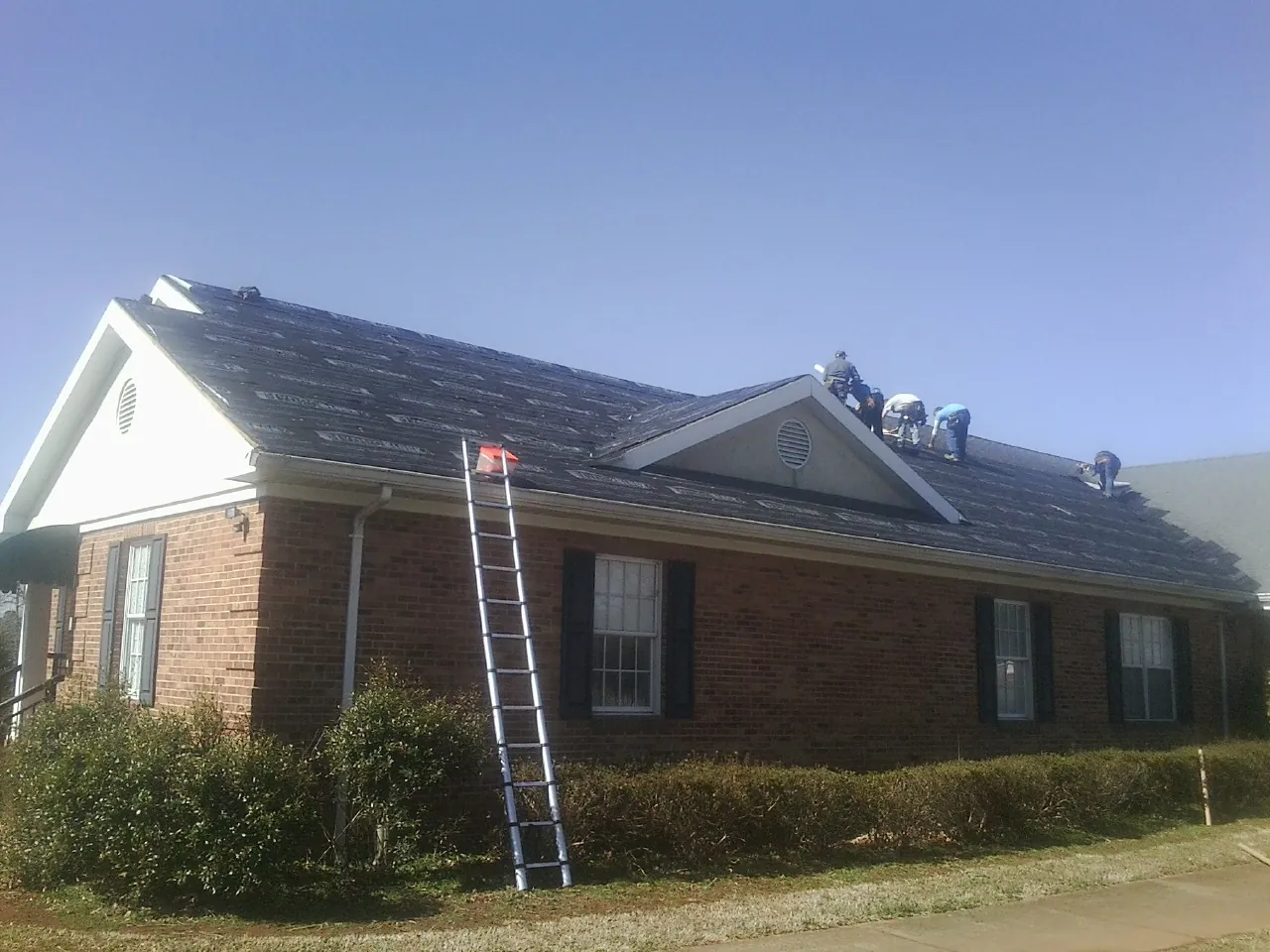 Crew tearing shingles off a brick ranch roof with a ladder set — Emergency Roof Repair work by our crew in Eliot
