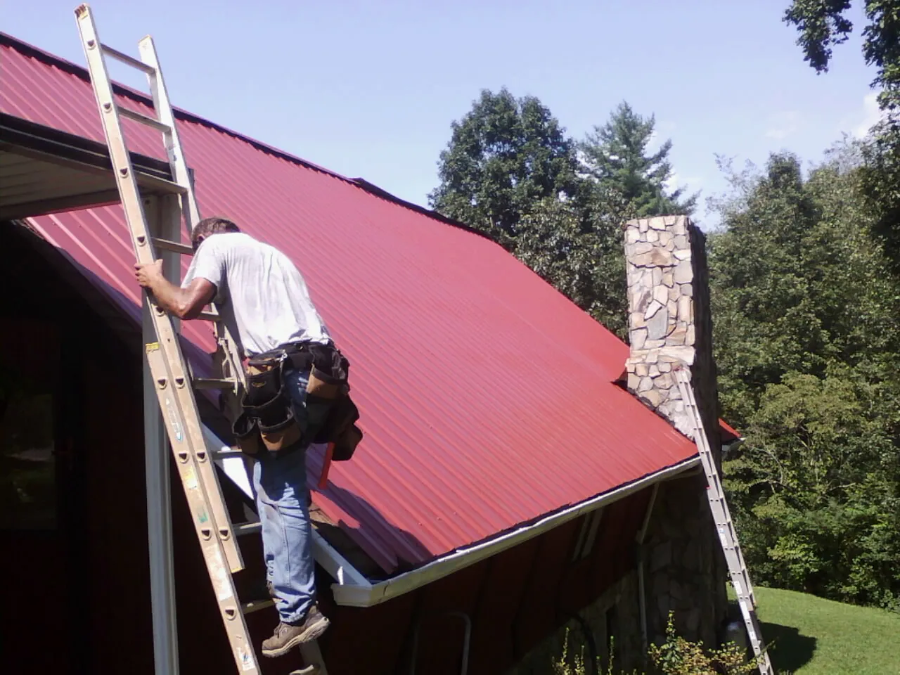 Roofer on a ladder beside a red metal roof and stone chimney — Soffit & Fascia Repair by Roofing Brothers in Eliot
