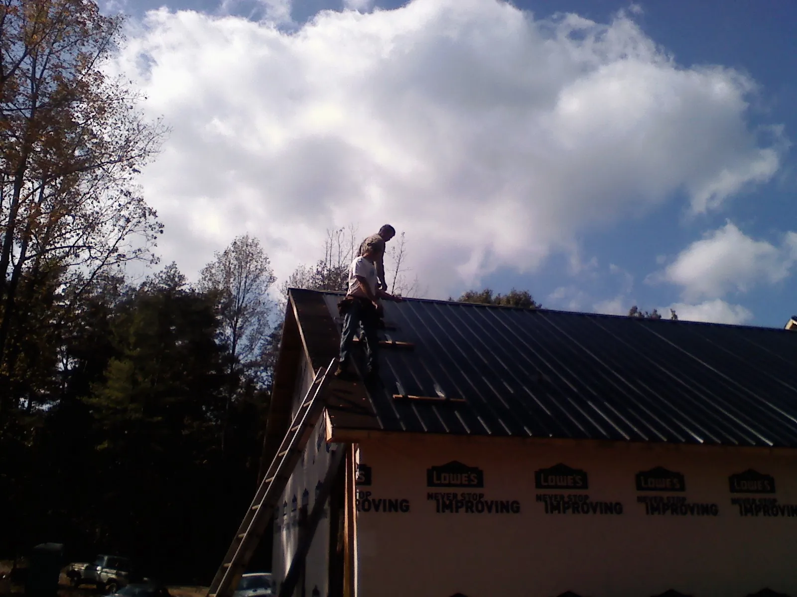 Roofer fastening dark metal panels high against a cloudy sky, from a Roof Inspection job in Eliot